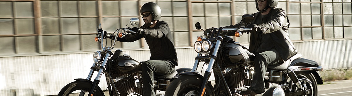Two men in all black protective gear riding black Harley® motorcycles near an industrial warehouse.